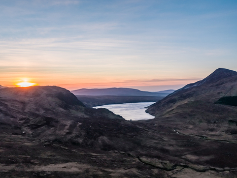 Aerial view of Altan Lough nestled among the rugged hills of County Donegal at sunset with golden light reflecting on the water and dark undulating terrain County Donegal Ireland.