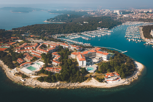 Aerial view of a luxurious resort nestled on a rocky peninsula where turquoise waters meet lush greenery and a bustling marina Pula Istria County Croatia.