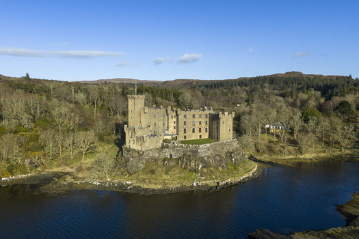Aerial view of Dunvegan Castle and Gardens situated on a rocky outcrop surrounded by dense forest and calm water under a clear blue sky Isle of Skye Scotland United Kingdom. Aerial view of Dunvegan Castle and Gardens situated on a rocky outcrop surrounded by dense forest and calm water under a clear blue sky Isle of Skye Scotland United Kingdom.
