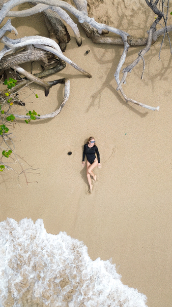 Aerial view of a woman in a black swimsuit lying on the sand at Grays Beach surrounded by driftwood and ocean waves in Honolulu Hawaii United States.