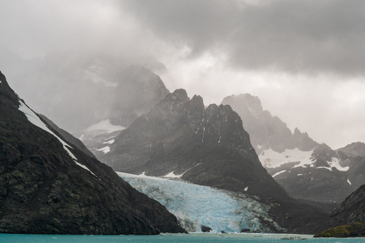 View of Drygalski Fjord featuring dramatic snow capped mountains a blue glacier tongue descending into icy water and a moody overcast sky in South Georgia and the South Sandwich Islands.