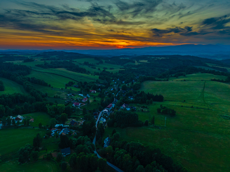 Aerial view of the village of Horni Zdar surrounded by lush green fields and dense forests under a vibrant sunset sky with orange clouds Jindrichuv Hradec 1 South Bohemian Region Czechia.