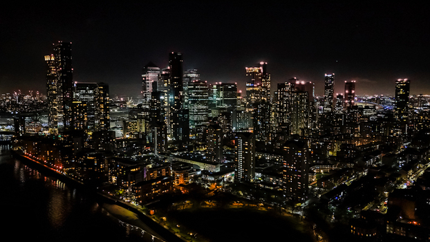Aerial view of the illuminated Canary Wharf skyline at night with modern skyscrapers and glowing windows reflecting over the dark River Thames in London England United Kingdom.