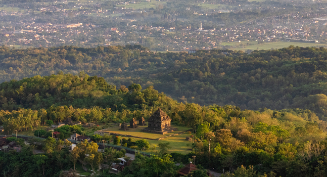 Aerial view of Ijo Temple nestled in lush forest under golden hour light with distant urban landscape in Kecamatan Prambanan Daerah Istimewa Yogya Indonesia.