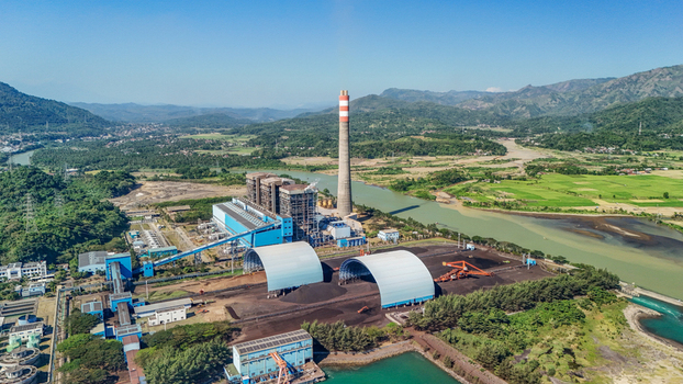 Aerial view of a power plant with coal storage domes situated along a river in a lush landscape in Indonesia.