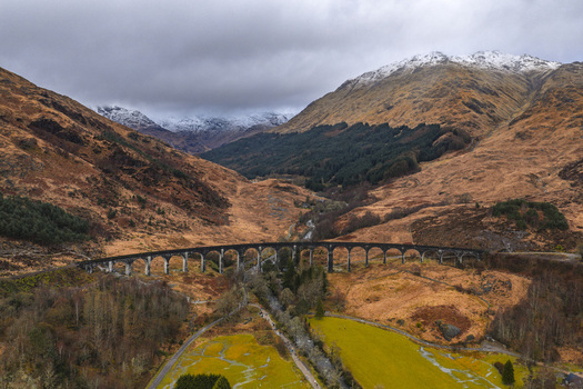 Aerial view of the historic Glenfinnan Viaduct with its curved concrete arches spanning a rugged valley under snow capped mountains Glenfinnan Scotland United Kingdom.