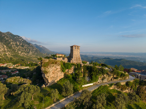 Aerial view of the historic Kruje Castle watchtower perched on a rocky cliff with winding roads and mountain peaks under a clear blue sky in Kruje Durres County Albania.