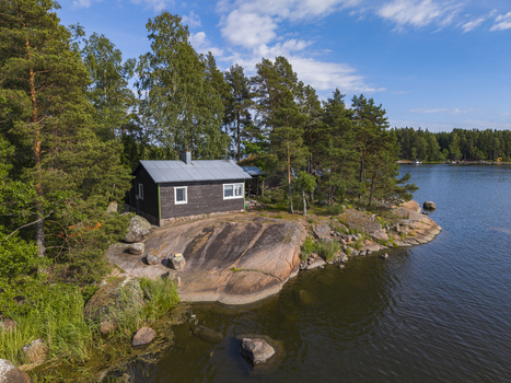 Aerial view of a small wooden cabin nestled on a rocky shoreline surrounded by pine trees and calm lake waters in Hamina Kymenlaakso Finland. Aerial view of a small wooden cabin nestled on a rocky shoreline surrounded by pine trees and calm lake waters in Hamina Kymenlaakso Finland.