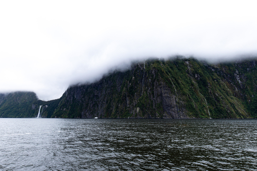 View of cascading waterfalls spilling from misty verdant cliffs into the dark rippled waters of the fjord a serene landscape under a cloudy sky Milford Sound New Zealand.