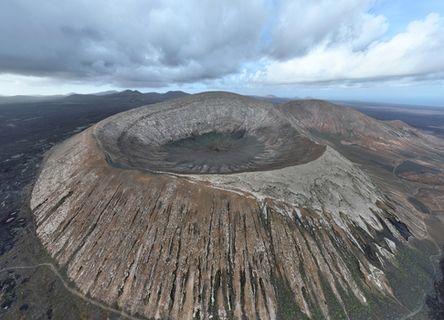 Aerial view of the rugged volcanic crater of Caldera Blanca with dark rocky slopes and deep interior basin under a cloudy sky in Lanzarote. Aerial view of the rugged volcanic crater of Caldera Blanca with dark rocky slopes and deep interior basin under a cloudy sky in Lanzarote.