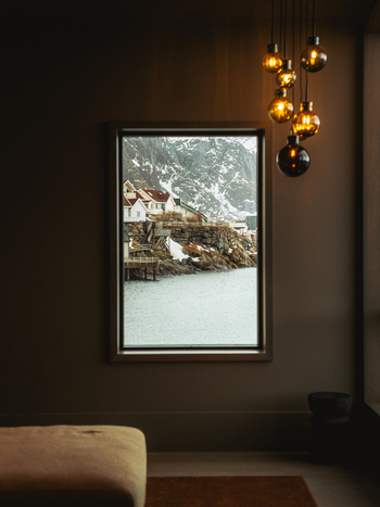 View of a framed window revealing a serene coastal village nestled amidst snow dusted cliffs and tranquil waters Henningsv&aelig;r Nordland Norway.