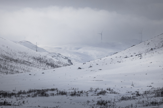View of a snowy mountainside with wind turbines emerging from thick fog under a cloudy sky in Ersfjord Troms Norway. View of a snowy mountainside with wind turbines emerging from thick fog under a cloudy sky in Ersfjord Troms Norway.