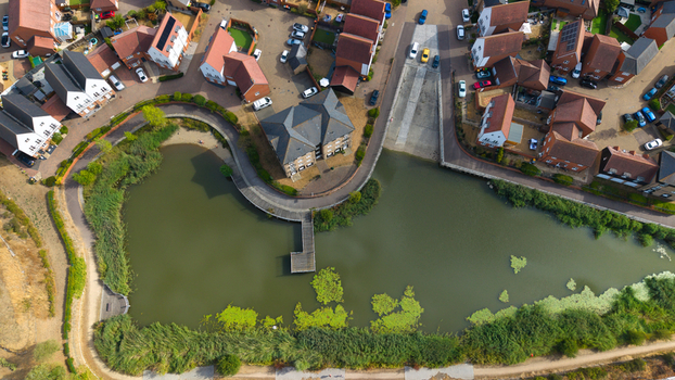 Aerial view of a modern residential housing estate featuring a central pond with a wooden walkway surrounded by houses with red tiled roofs in Bedford England United Kingdom. Aerial view of a modern residential housing estate featuring a central pond with a wooden walkway surrounded by houses with red tiled roofs in Bedford England United Kingdom.