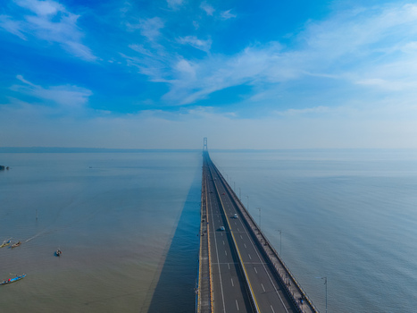 Aerial view of the Suramadu Bridge stretching across the Madura Strait with small fishing boats below under a clear blue sky in Kecamatan Kenjeran Jawa Timur Indonesia.