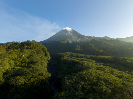 Aerial view of the majestic Mount Merapi volcano with steam rising from its crater above a dense tropical forest under a clear blue sky in Kecamatan Pakem Daerah Istimewa Yogya Indonesia. Aerial view of the majestic Mount Merapi volcano with steam rising from its crater above a dense tropical forest under a clear blue sky in Kecamatan Pakem Daerah Istimewa Yogya Indonesia.