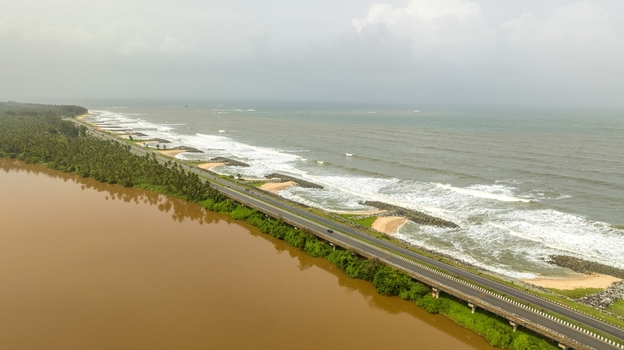Aerial view of Maravanthe Beach featuring the scenic highway running between the Arabian Sea and the Suparnika River lined with lush palm trees in Maravanthe Karnataka India. Aerial view of Maravanthe Beach featuring the scenic highway running between the Arabian Sea and the Suparnika River lined with lush palm trees in Maravanthe Karnataka India.