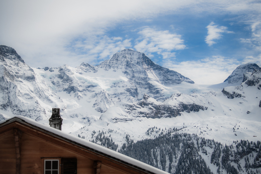 View of snow covered mountains piercing the blue sky contrasting with the warm tones of a building s roof in the foreground Murren Switzerland. View of snow covered mountains piercing the blue sky contrasting with the warm tones of a building s roof in the foreground Murren Switzerland.