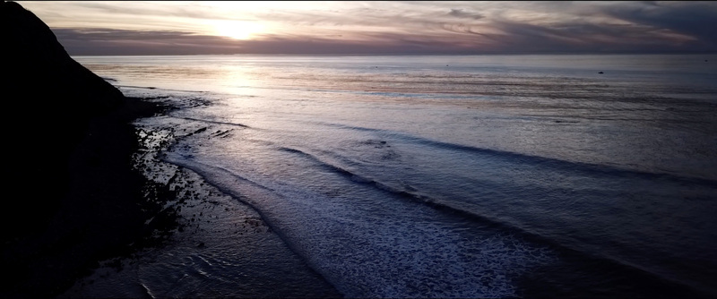 Aerial view of the Pacific Ocean coastline at sunset with gentle waves rolling onto the rocky shore under a soft twilight sky in Los Angeles California United States.