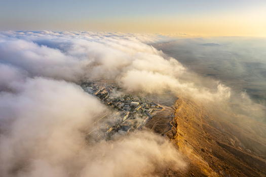 Aerial view of Mitzpe Ramon town perched on the edge of the Makhtesh Ramon crater partially obscured by thick clouds during the golden hour in Mitzpe Ramon South District Israel. Aerial view of Mitzpe Ramon town perched on the edge of the Makhtesh Ramon crater partially obscured by thick clouds during the golden hour in Mitzpe Ramon South District Israel.