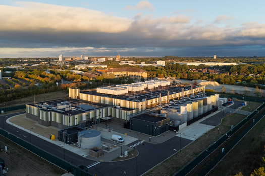 Aerial view of a modern data center facility with industrial architecture rooftop cooling units and a perimeter fence set against trees and a distant town in United Kingdom. Aerial view of a modern data center facility with industrial architecture rooftop cooling units and a perimeter fence set against trees and a distant town in United Kingdom.