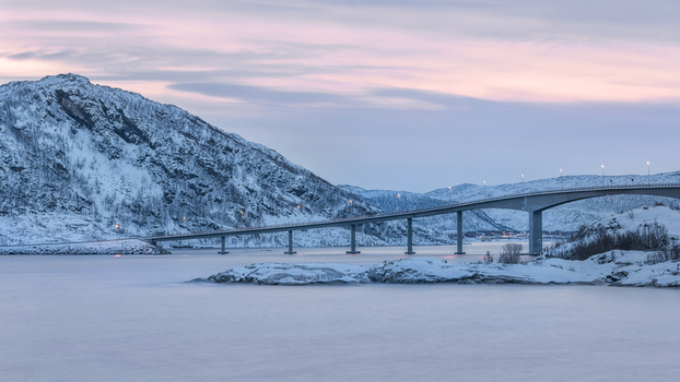View of a long bridge spanning across a fjord with snow covered mountains under a soft pink twilight sky Sommaroy Troms Norway. View of a long bridge spanning across a fjord with snow covered mountains under a soft pink twilight sky Sommaroy Troms Norway.