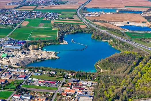 Aerial view of the bright turquoise lake reflecting the sky contrasting with the surrounding green fields and industrial areas Sausheim Grand Est France.