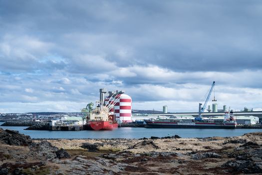 Hafnarfjordur Iceland 17 April 2026 View of the industrial port with cargo ships a red and white striped storage tank and cranes under a cloudy sky.