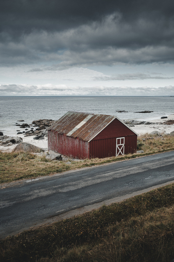 View of a traditional red wooden cabin at Vikten Beach with a rusty roof and white door frame situated by the rocky coastline and dark ocean in Vikten Nordland Norway.