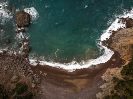 Aerial view of the rugged rocky coastline of Calafuria with turquoise waves crashing against the dark sandy beach and steep cliffs Calafuria Toscana Italy.