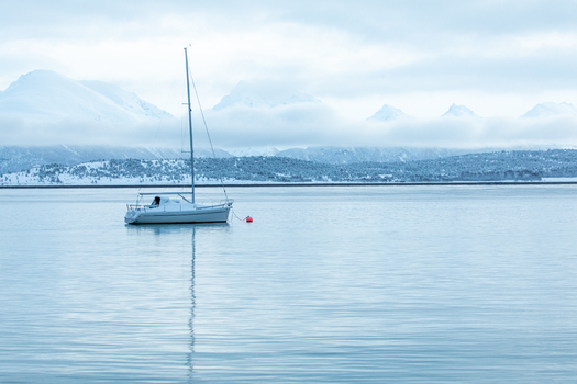 View of a white sailboat anchored on calm water with snow covered mountains and low clouds in the background Tromso Troms Norway. View of a white sailboat anchored on calm water with snow covered mountains and low clouds in the background Tromso Troms Norway.