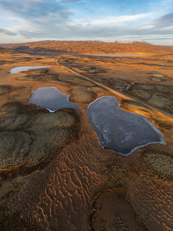 Aerial view of the volcanic landscape with mossy terrain small ponds and a road winding through the rugged terrain under a golden hour sky in Reykjanesbaer Iceland. Aerial view of the volcanic landscape with mossy terrain small ponds and a road winding through the rugged terrain under a golden hour sky in Reykjanesbaer Iceland.