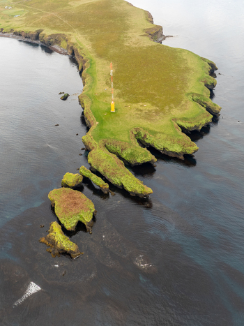 Aerial view of the Tjornes peninsula featuring green cliffs rocky coastal formations and a yellow navigation tower surrounded by dark ocean waters in Tjorneshreppur Iceland. Aerial view of the Tjornes peninsula featuring green cliffs rocky coastal formations and a yellow navigation tower surrounded by dark ocean waters in Tjorneshreppur Iceland.