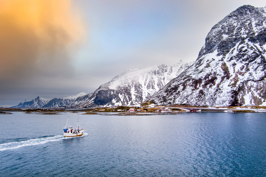 Aerial view of a fishing boat navigating the calm waters of a fjord in Lofoten surrounded by snow capped mountains under a dramatic sky in Nordland Norway.