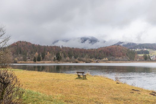 View of a wooden bench on the grassy shore of a calm lake with autumn trees and misty mountains in Hallstatt Upper Austria Austria. View of a wooden bench on the grassy shore of a calm lake with autumn trees and misty mountains in Hallstatt Upper Austria Austria.