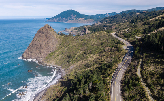 Aerial view of Highway 101 winding along the rugged coastline with steep cliffs dense forests and the Pacific Ocean in Port Orford Oregon United States. Aerial view of Highway 101 winding along the rugged coastline with steep cliffs dense forests and the Pacific Ocean in Port Orford Oregon United States.