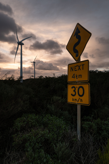 View of wind turbines at the Sandpatch Wind Farm with road signs indicating a winding road and speed limit in the foreground in Sandpatch Western Australia Australia. View of wind turbines at the Sandpatch Wind Farm with road signs indicating a winding road and speed limit in the foreground in Sandpatch Western Australia Australia.