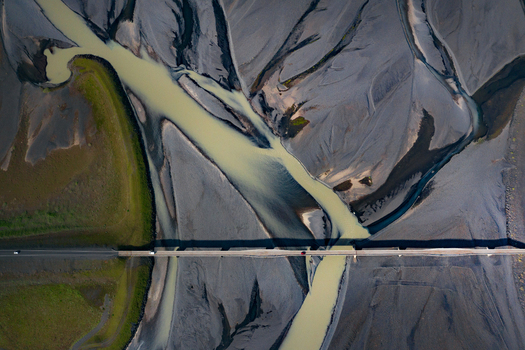 Aerial view of a braided glacial river with silty water flowing through dark volcanic sand and a bridge with cars crossing the stream in Iceland.