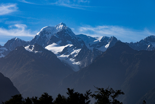 View of snow capped peaks pierce the azure sky casting long shadows across the dark forested slopes in Lake Matheson West Coast Region New Zealand. View of snow capped peaks pierce the azure sky casting long shadows across the dark forested slopes in Lake Matheson West Coast Region New Zealand.