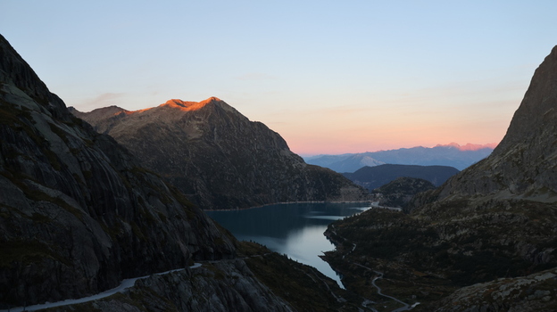 View of Bel Oiseau mountain peak reflecting in the calm waters of Emosson Lake at sunset with rugged rocky slopes and a winding mountain road in Finhaut Valais Switzerland. View of Bel Oiseau mountain peak reflecting in the calm waters of Emosson Lake at sunset with rugged rocky slopes and a winding mountain road in Finhaut Valais Switzerland.