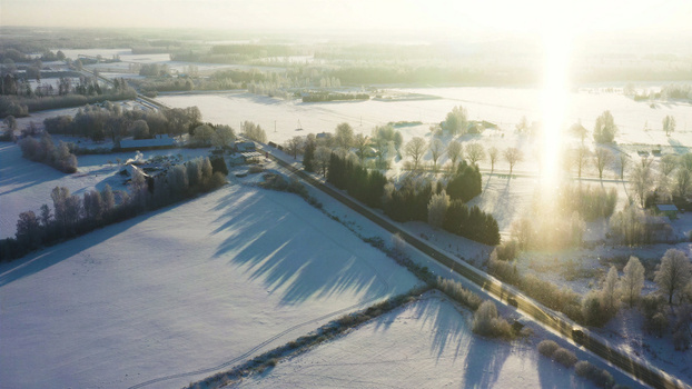 Aerial view of a snowy landscape with frost covered trees open fields and a road illuminated by a bright sun flare in Latvia. Aerial view of a snowy landscape with frost covered trees open fields and a road illuminated by a bright sun flare in Latvia.
