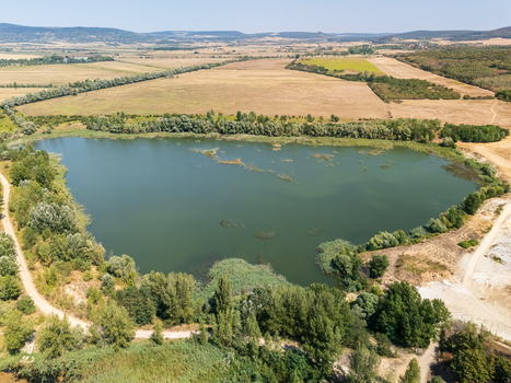 Aerial view of Burnout mining lake in Veszprem County Hungary with shoreline reeds forested hills and rural wetlands under a clear sunny sky.