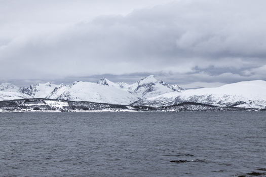 View of snow capped mountains and dark fjord waters under a cloudy sky in Tromso Troms Norway.