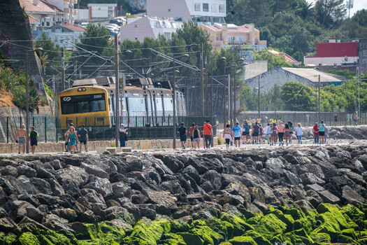 Cruz Quebrada Portugal 03 May 2026 View of the Passeio Maritimo de Alges with people walking along the waterfront as a yellow CP train passes by historic buildings and lush trees.