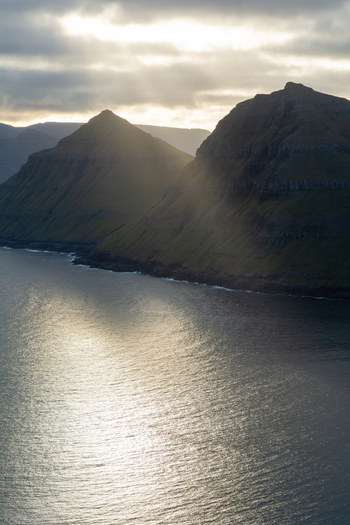 View of dramatic green mountains rising from the ocean with sunlight reflecting on the water surface Faroe Islands. View of dramatic green mountains rising from the ocean with sunlight reflecting on the water surface Faroe Islands.
