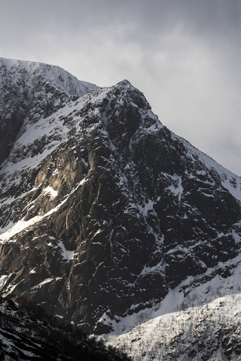 View of a steep snow covered mountain peak with dark rocky cliffs under an overcast sky in Kvaloya Troms Norway. View of a steep snow covered mountain peak with dark rocky cliffs under an overcast sky in Kvaloya Troms Norway.