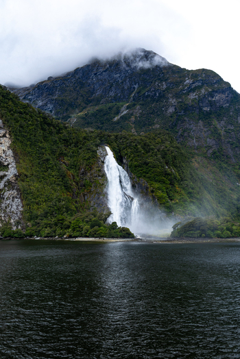 View of cascading waterfalls plummeting down the verdant steep mountainside into the dark reflective waters under a misty sky Milford Sound Southland Region New Zealand.
