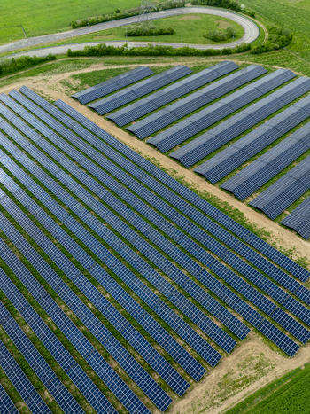 Aerial view of rows of solar panels in a large photovoltaic power station surrounded by green fields and a winding road.