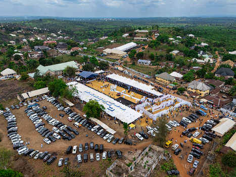 Aerial view of a vibrant outdoor gathering with white tents a golden central space and a dense array of vehicles nestled amidst green hills ohafia Abia Nigeria. Aerial view of a vibrant outdoor gathering with white tents a golden central space and a dense array of vehicles nestled amidst green hills ohafia Abia Nigeria.