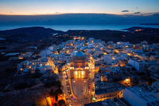Aerial view of the Parish Church of the Assumption of the Blessed Virgin Mary into Heaven illuminated at dusk surrounded by the town of L Imgarr Regjun Tramuntana Malta.