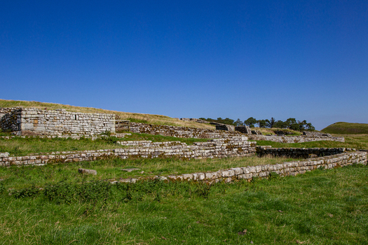 View of the ancient stone ruins of Vindolanda Roman Fort with low walls and grassy fields under a clear blue sky in Hexham England United Kingdom. View of the ancient stone ruins of Vindolanda Roman Fort with low walls and grassy fields under a clear blue sky in Hexham England United Kingdom.